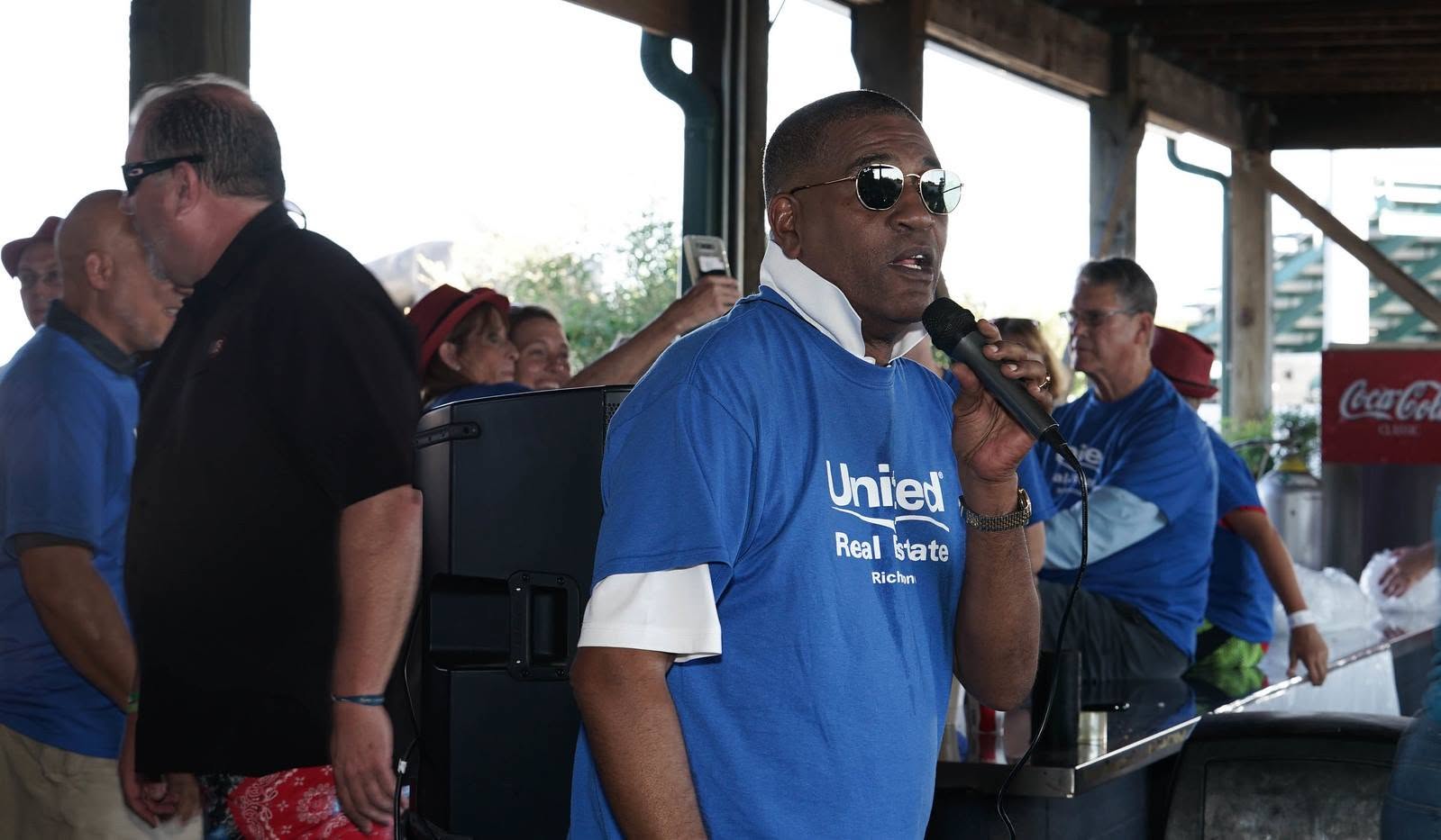 Person speaking into microphone at an outdoor event, wearing sunglasses and a blue shirt, with people in the background.