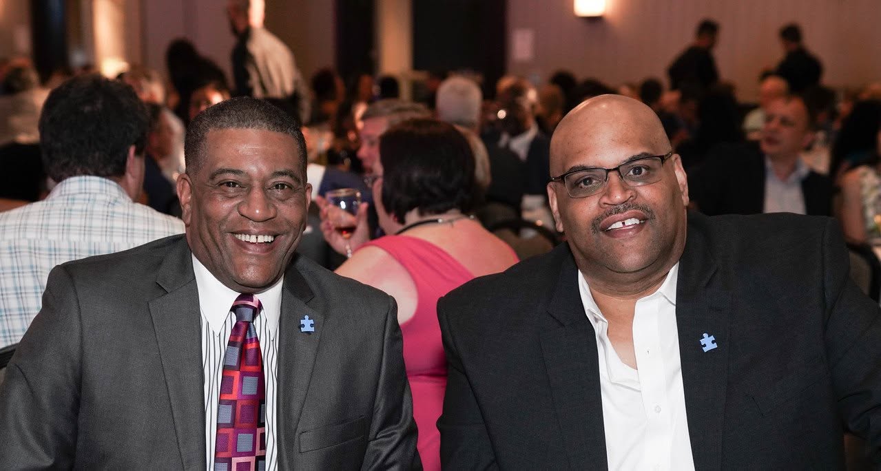 Two men in suits sitting at a table in a crowded event, smiling, with people in the background.