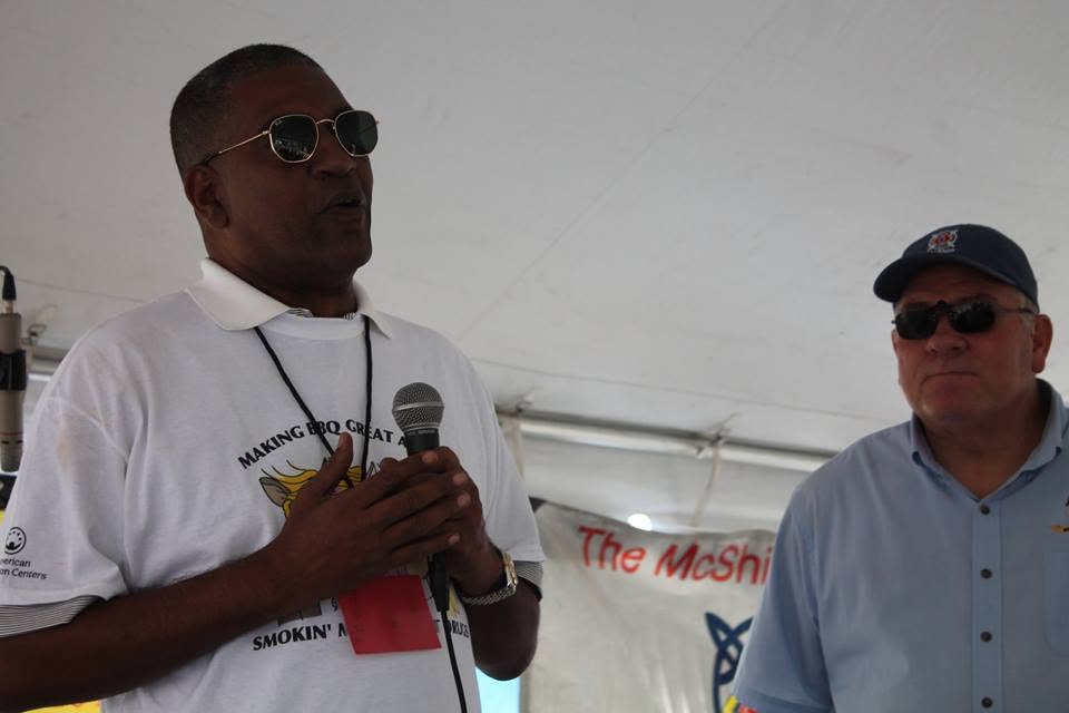 Two men standing under a tent, one holding a microphone, the other wearing sunglasses and a cap, with a banner in the background.