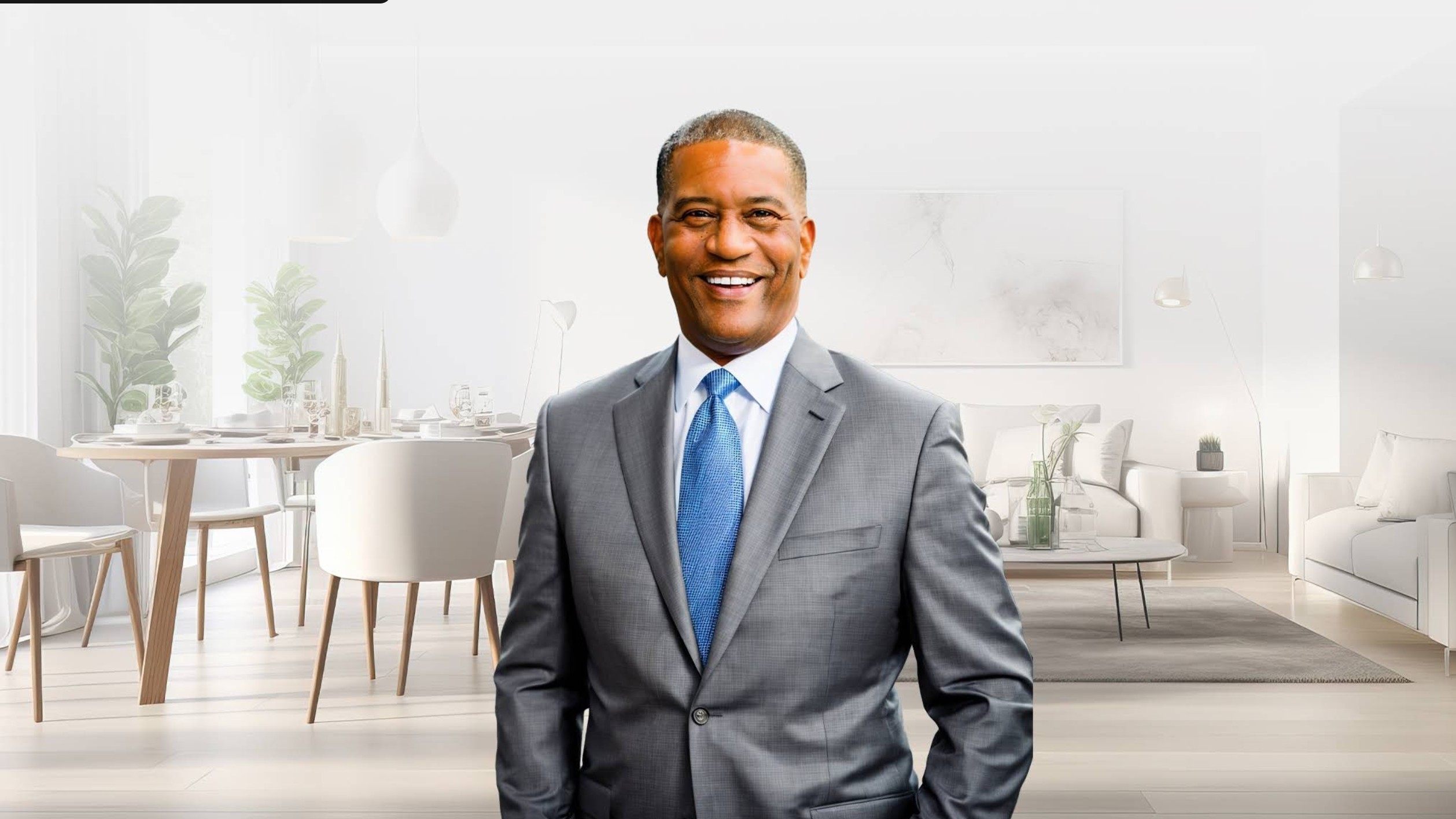 Smiling man in a suit standing in a modern, bright office or lounge area with chairs and tables.