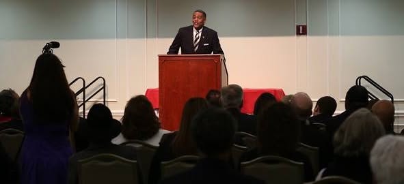 Man in suit speaking at podium in front of audience in a conference room.