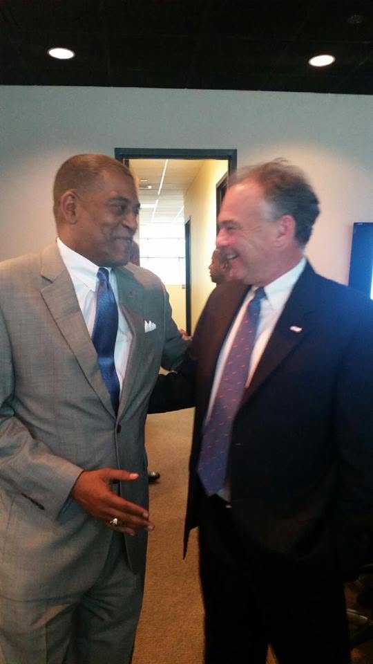 Two men in suits talking and smiling indoors, one with a gray suit and the other with a dark suit, in a professional setting.