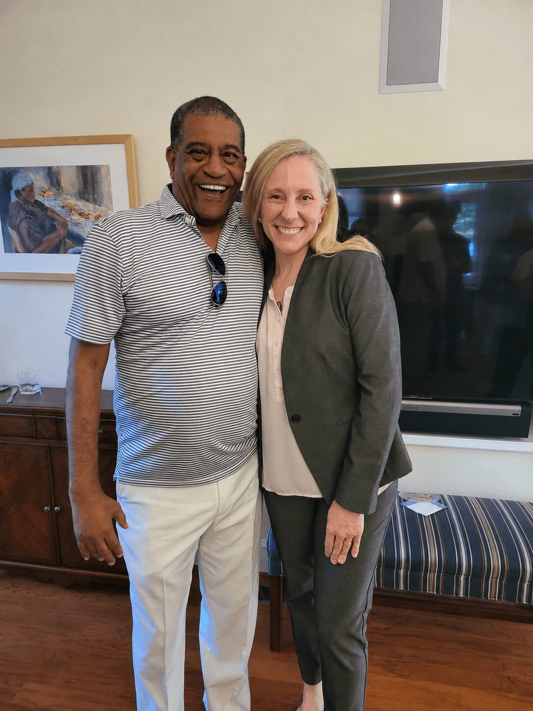 Two people, a man and a woman, stand close together smiling indoors, with a television and framed picture behind them.