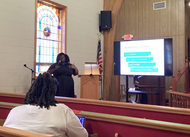 Woman presenting in a church with a stained glass window, American flag, and large screen displaying a presentation.