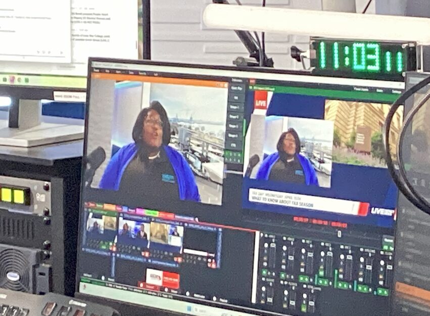 Television studio control room with monitors showing a woman speaking, and a digital clock displaying 11:03,11.