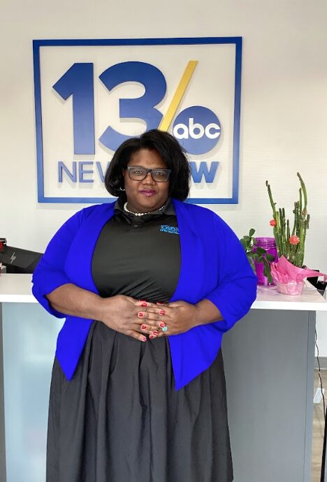 Woman with glasses and dark hair, wearing a black top and blue cardigan, standing in front of a news station sign, with flowers nearby.