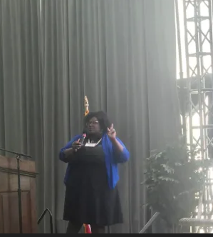 Person standing on stage with a curtain background, making peace signs with both hands, wearing a black dress and blue jacket.