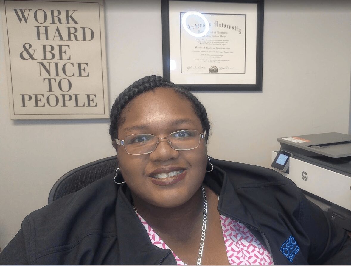 Woman with glasses and earrings smiling in an office, framed certificates on the wall behind her.