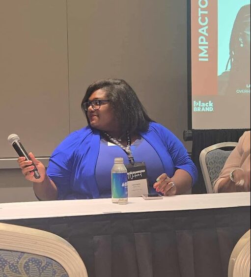 Woman with glasses and black hair, wearing a blue blazer, sitting at a table with a water bottle, holding a microphone.