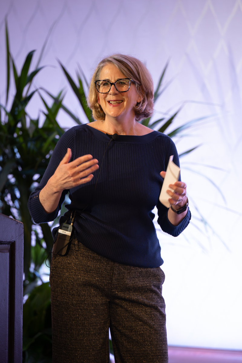 Woman with glasses smiling, holding a paper, gesturing with her right hand, standing in front of a light background with plants.