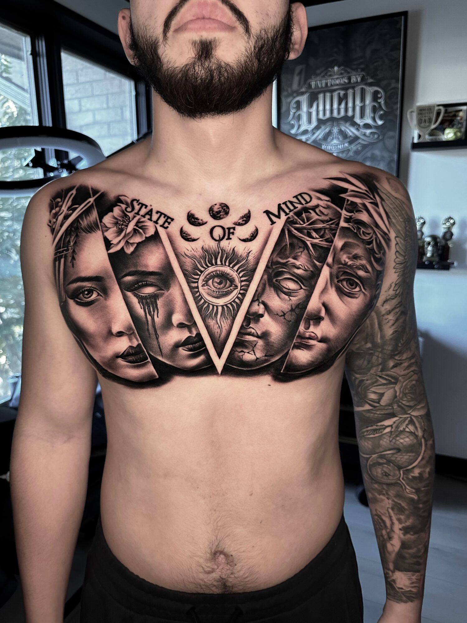 Tattooed chest with faces, eye, and text, in a tattoo studio setting, with a man showing his upper body.