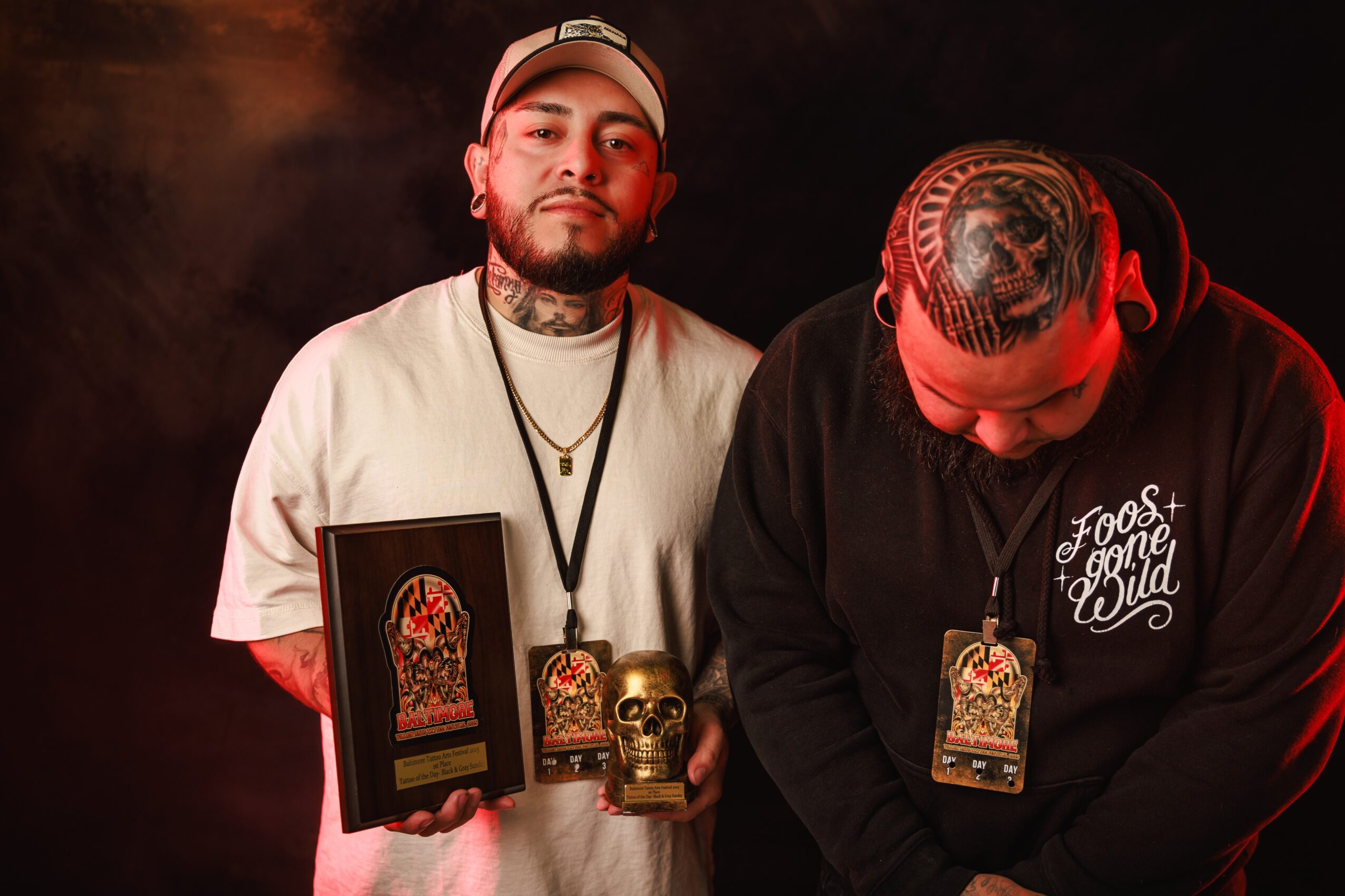 Two men stand against a dark background, one holding a framed artwork and a skull, the other looking down, wearing a hoodie.