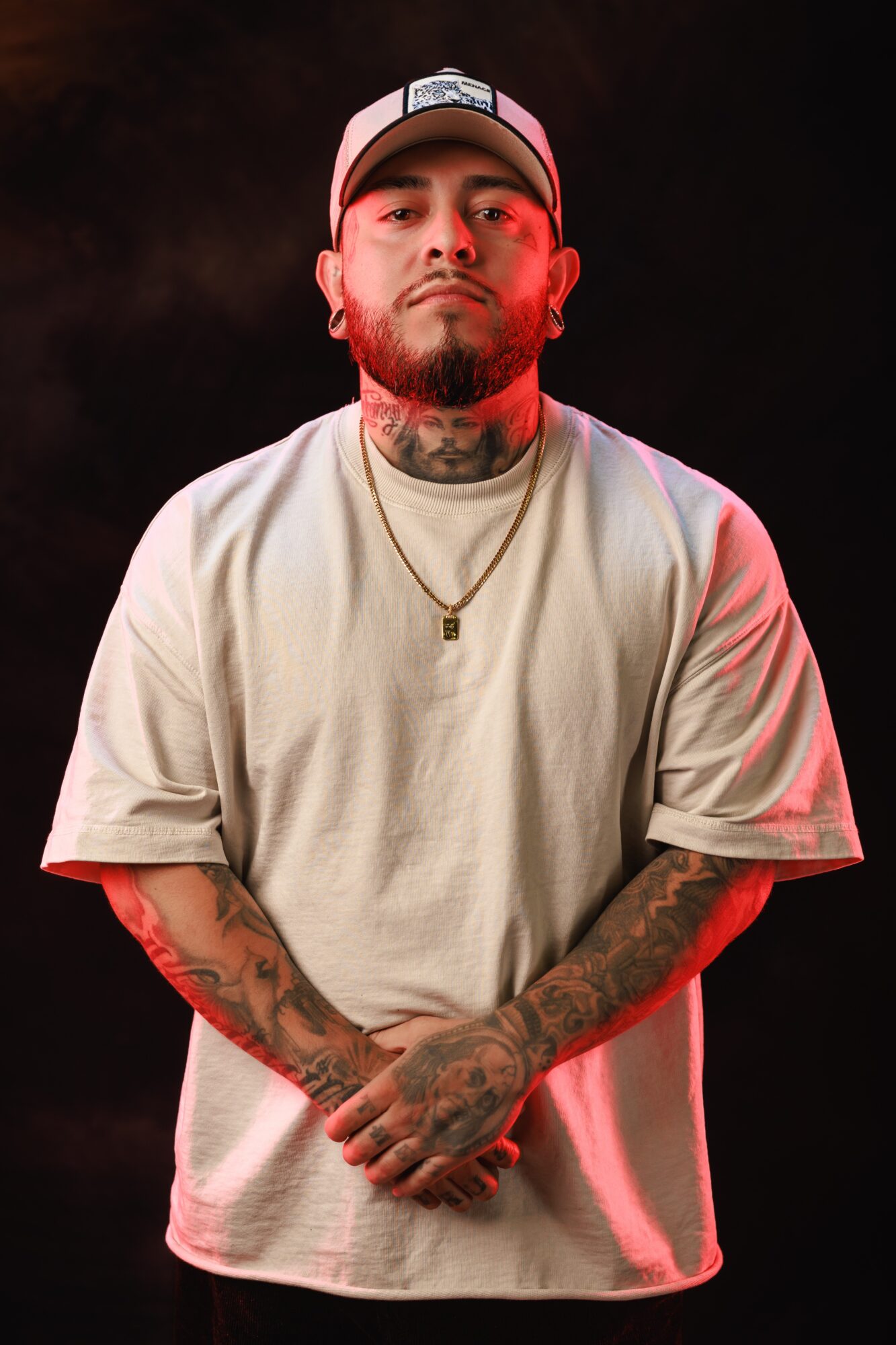 Man with tattoos, beard, and cap, wearing a light-colored T-shirt, standing against a dark background.
