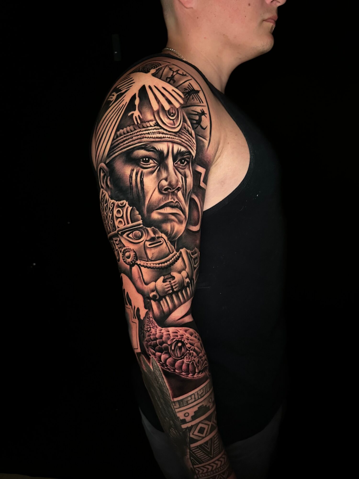 Tattooed arm with detailed face, headdress, and geometric patterns, visible from the side against a black background.