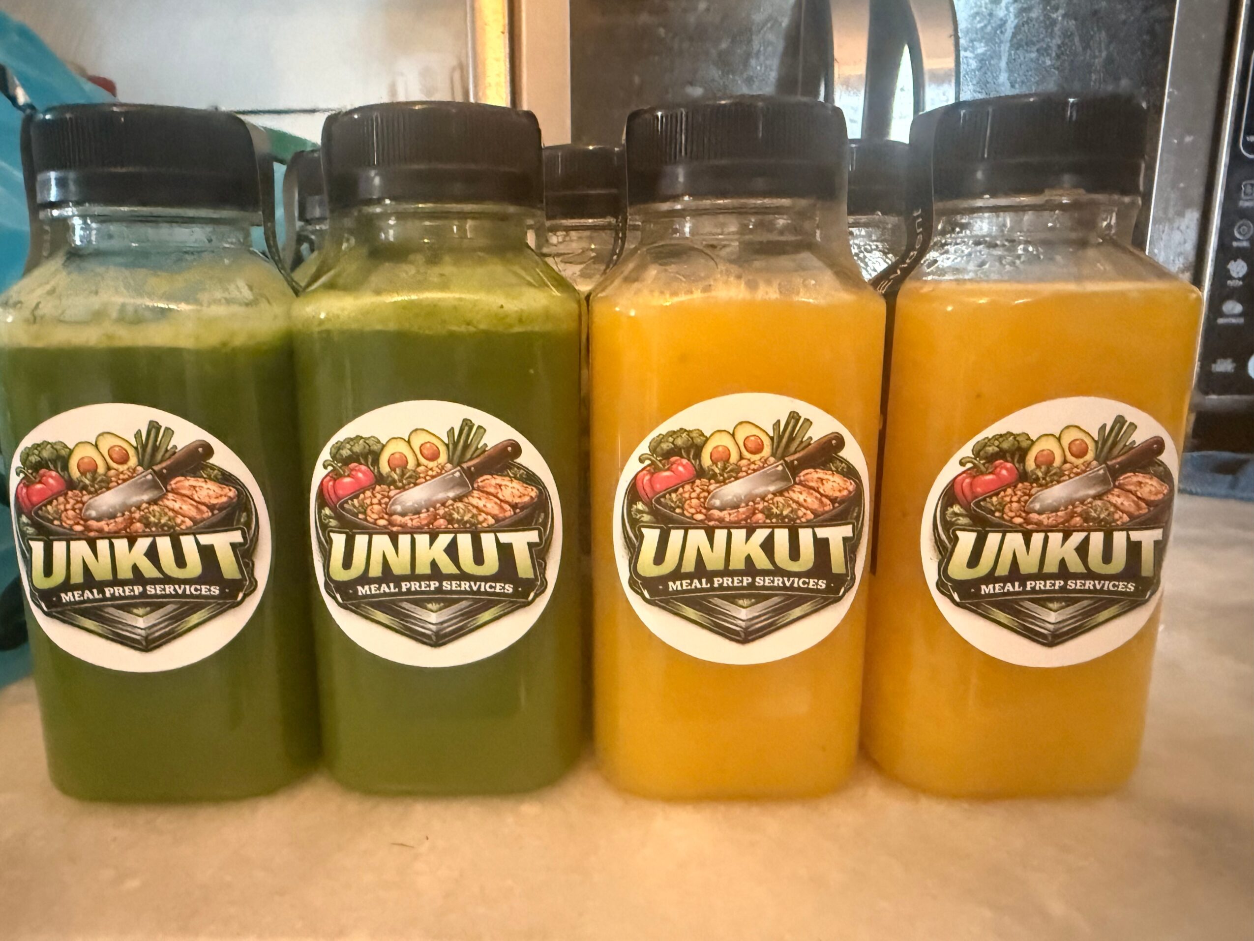 Four bottles of juice with black caps and labels, green and orange colors, on a beige surface.