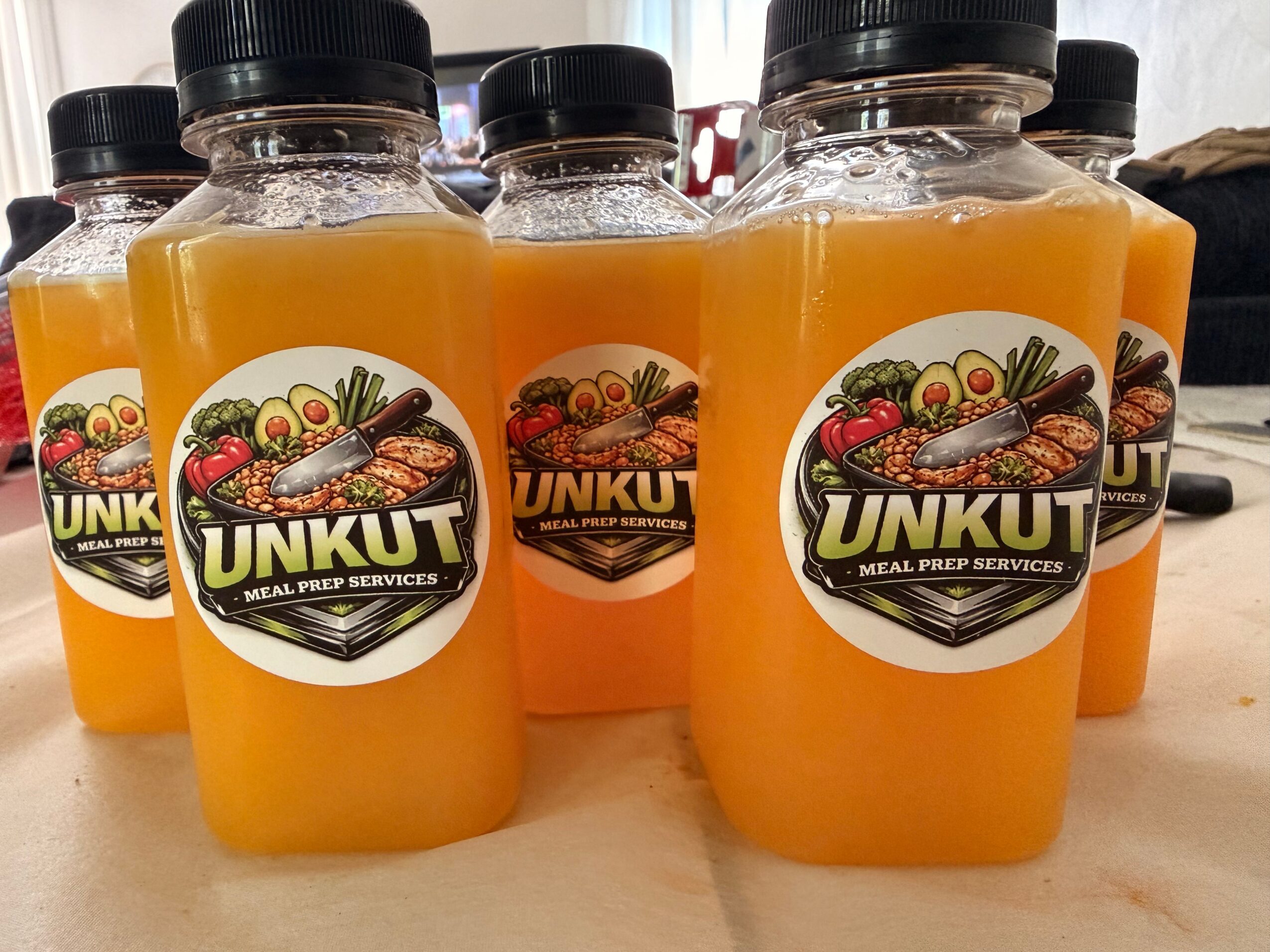 Six bottles of orange juice with black caps and UNKUT labels on a table.