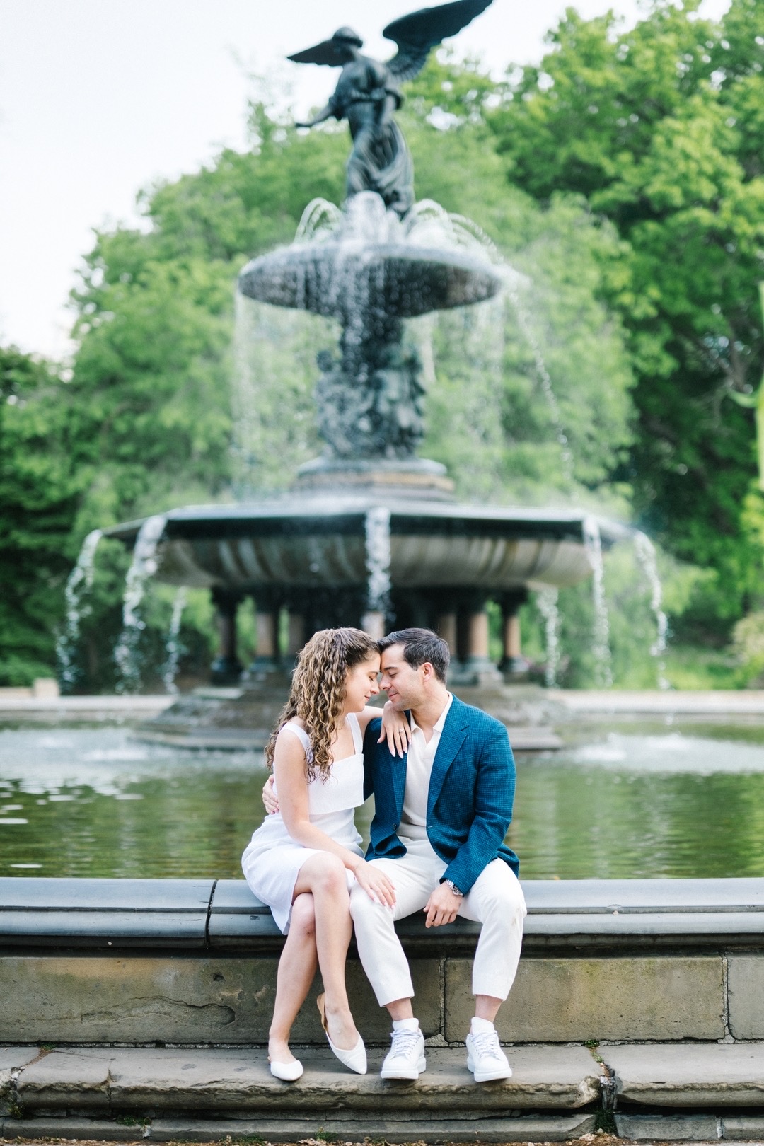 A couple sitting on a fountain's edge, with water flowing and a statue of a woman with wings above, surrounded by trees.