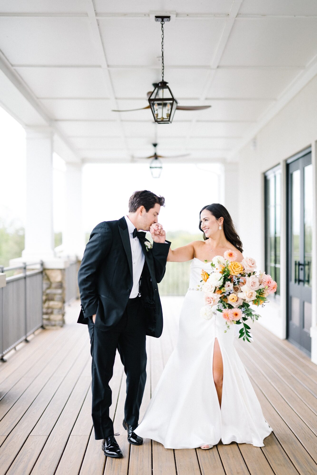 Bride and groom on porch, bride touching groom's face, bride holding bouquet, bright background, wedding attire, wooden floor