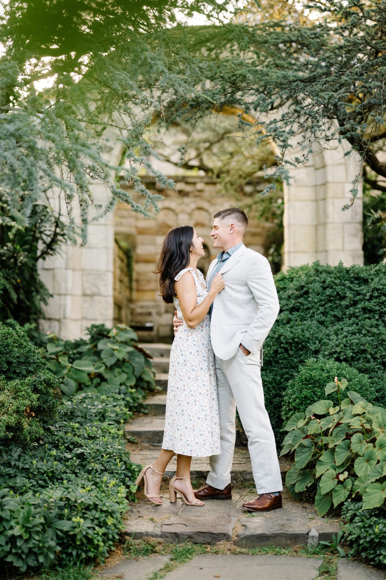 A couple stands close together in a garden, surrounded by greenery and stone structures, gazing into each other's eyes.