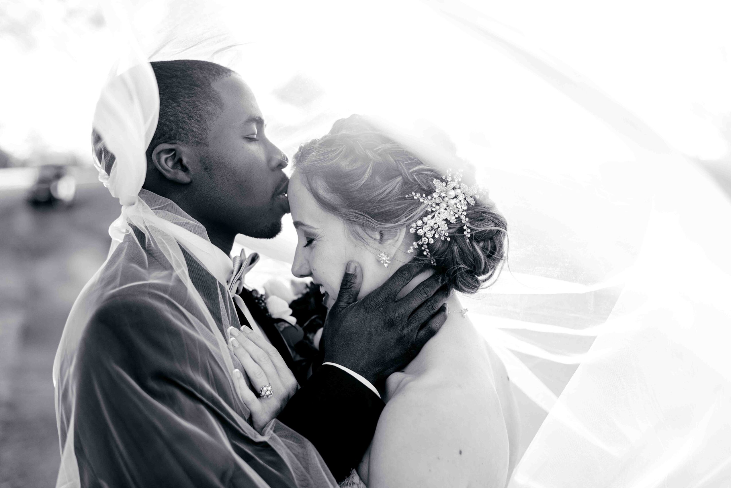 A couple in wedding attire share an intimate moment, with the man gently holding the woman's face and kissing her forehead.