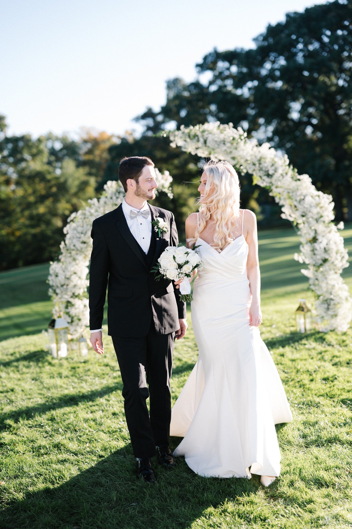 A bride and groom stand outdoors under a floral arch, holding hands, with trees and grass in the background.