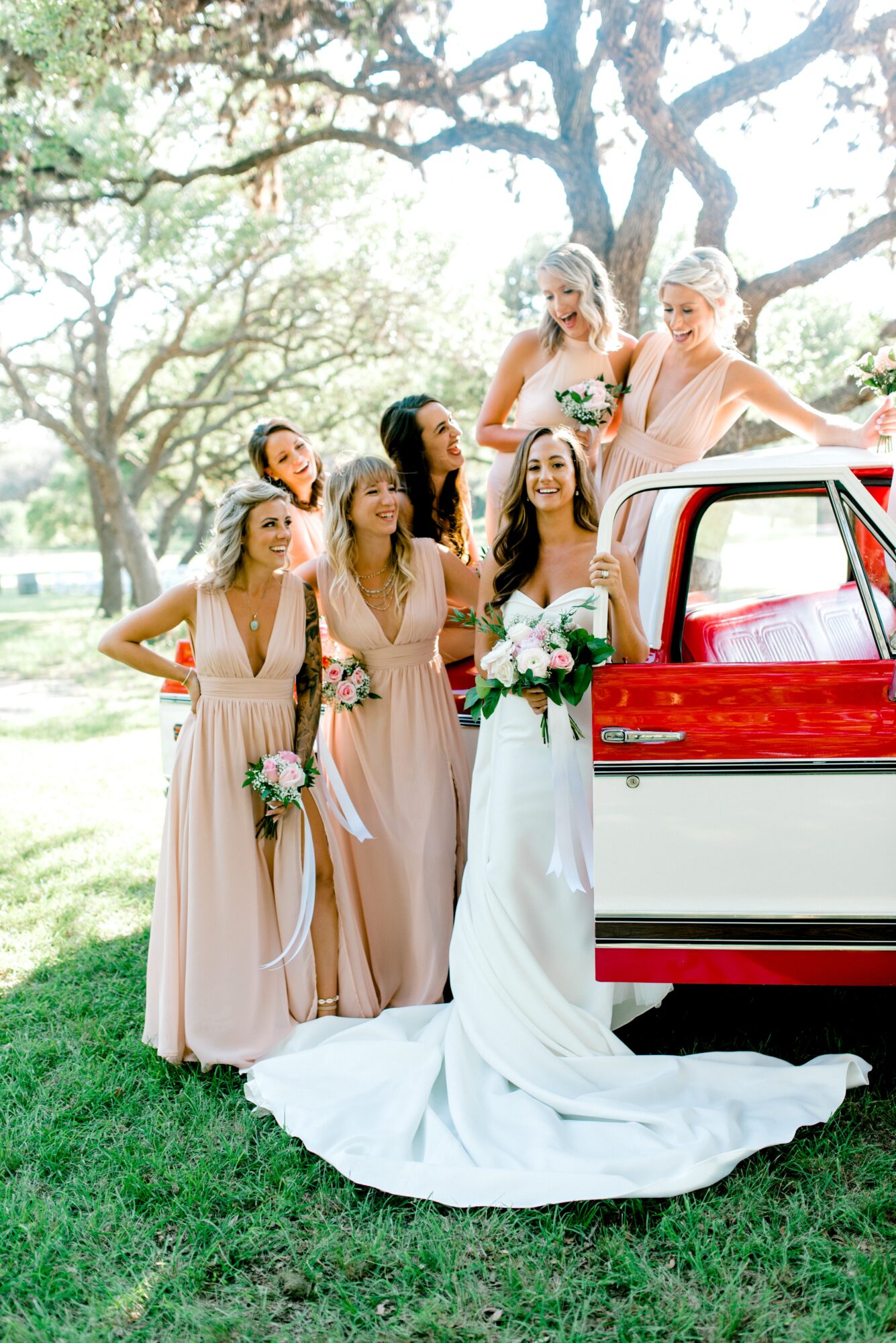 Group of women in dresses, one in wedding gown, standing outdoors near a red car, with trees and sunlight in background.