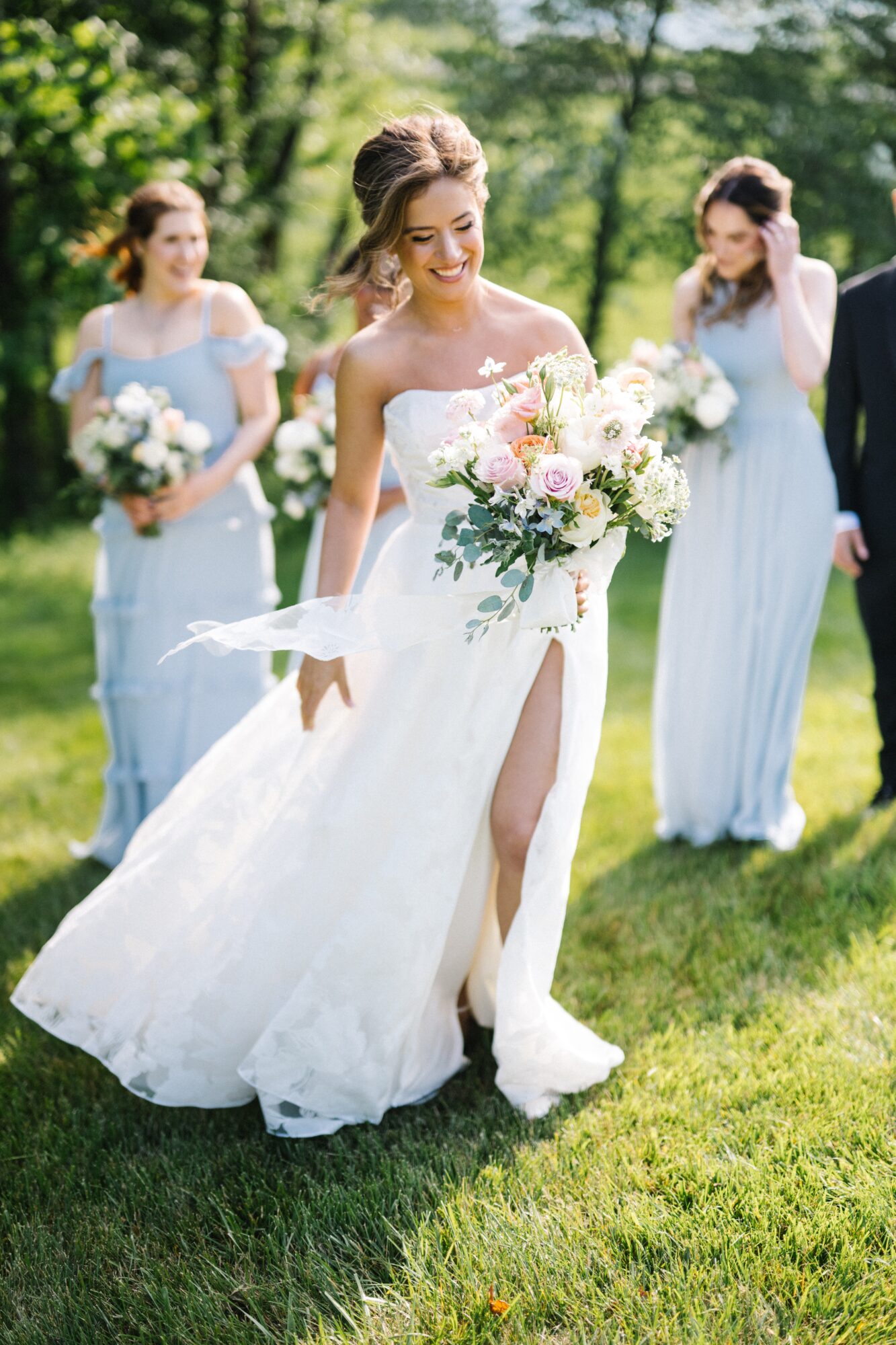 Bride in a white wedding dress holding a bouquet, smiling outdoors with three bridesmaids in light blue dresses in the background.