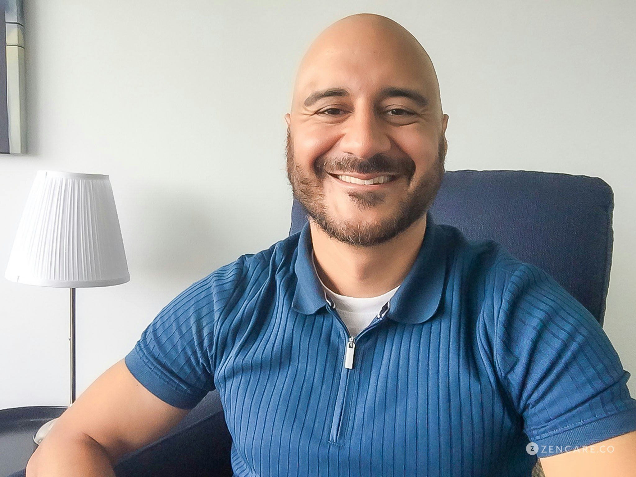 A smiling man with a beard and bald head sitting in a chair, wearing a blue shirt, in a room with a lamp and white wall.
