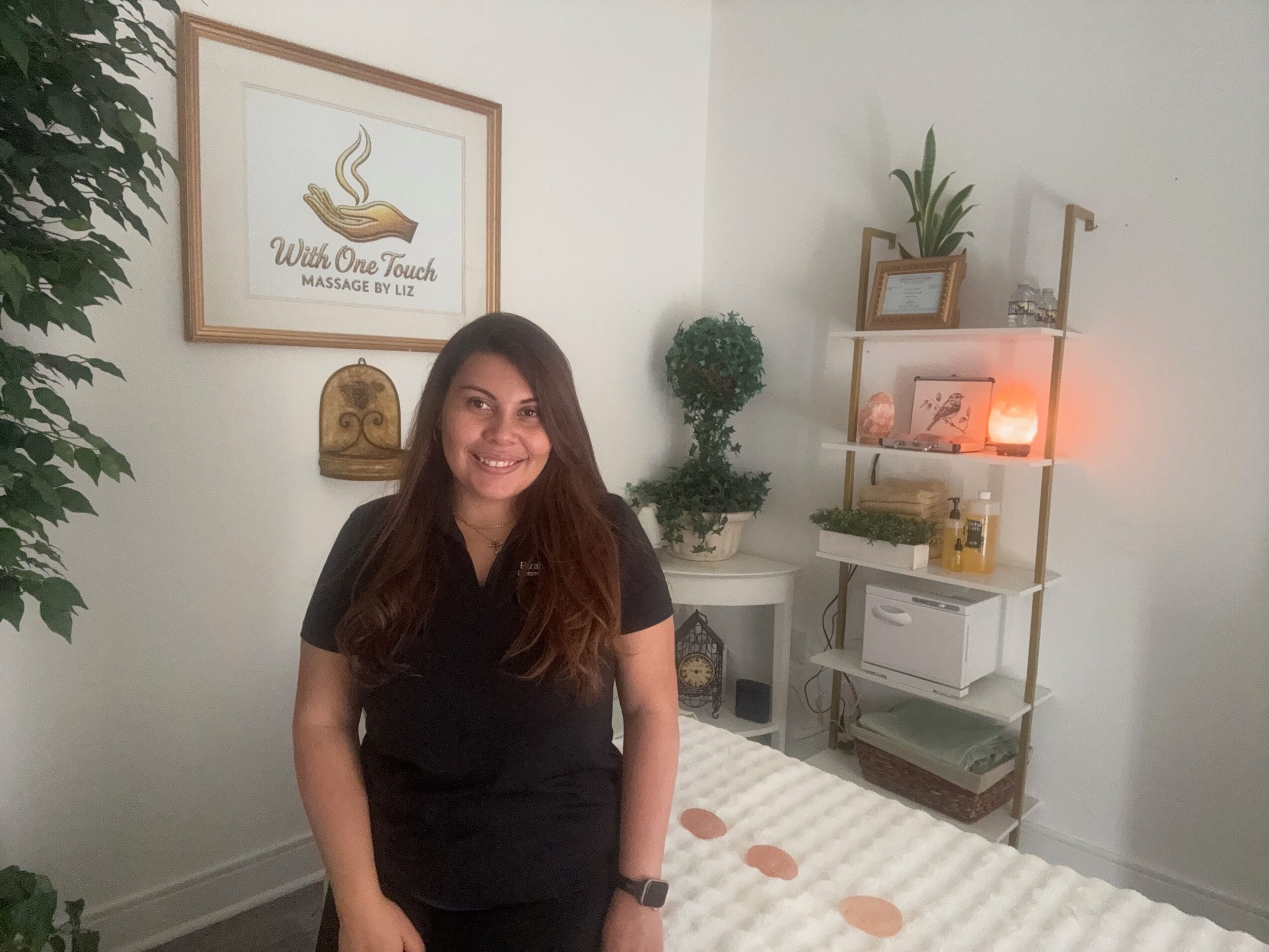 Woman with long hair smiling, standing in a room with shelves, plants, and framed artwork on the wall.
