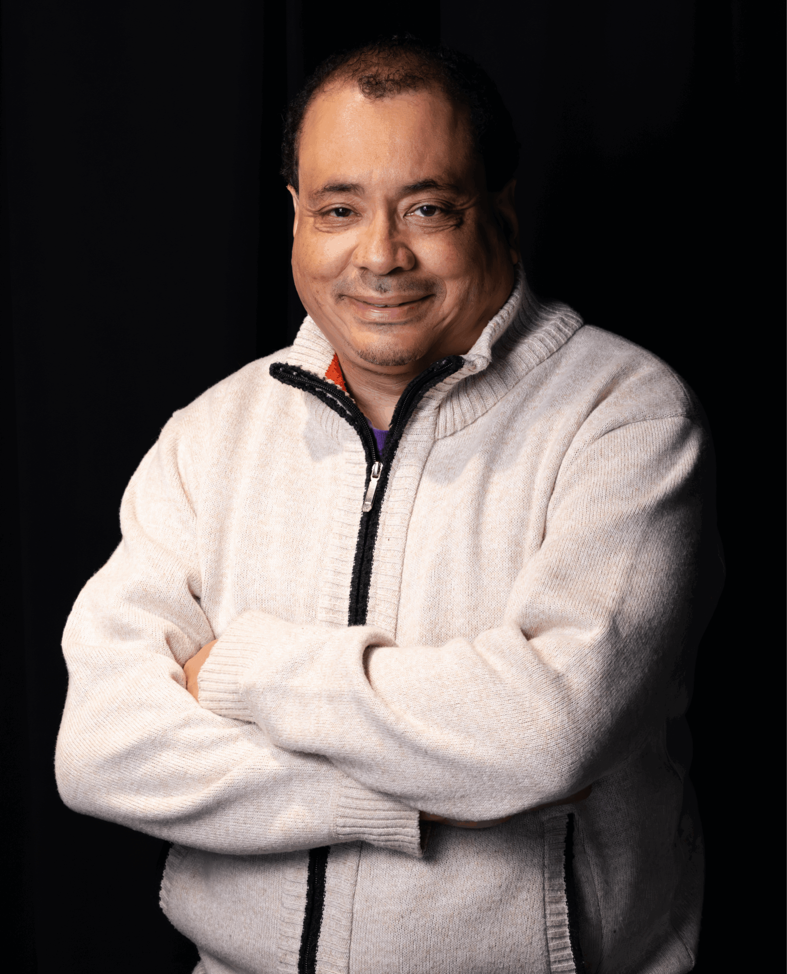 Man with crossed arms smiling, wearing a light-colored zip-up sweater, against a dark background.
