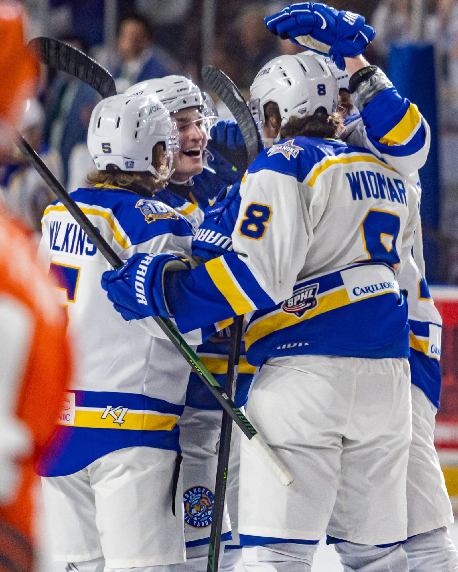 Hockey players in white and blue uniforms celebrating on the ice, some with helmets and sticks, in an arena.
