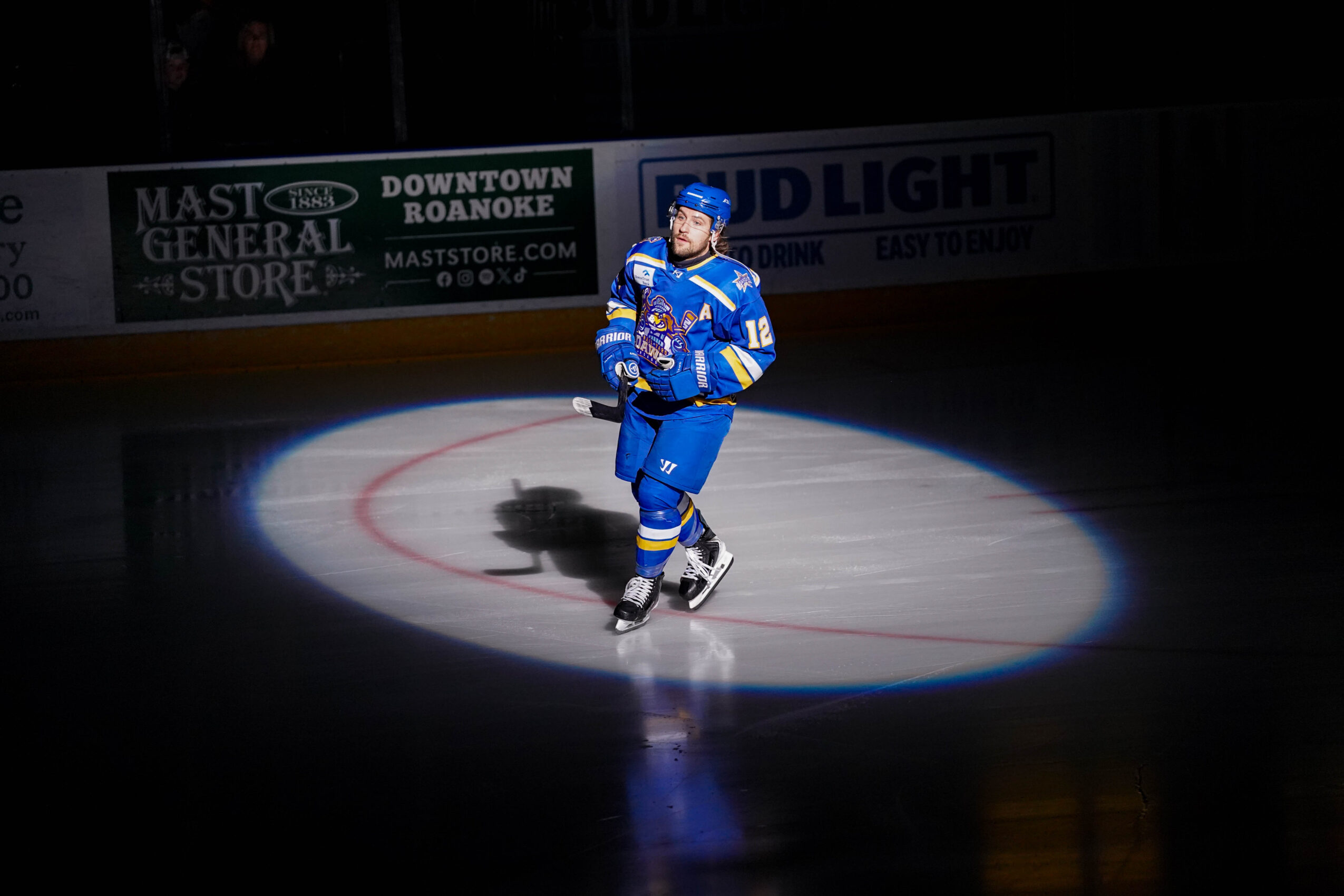 Hockey player in blue uniform skating on ice illuminated by spotlight, with dark background and advertising banners.