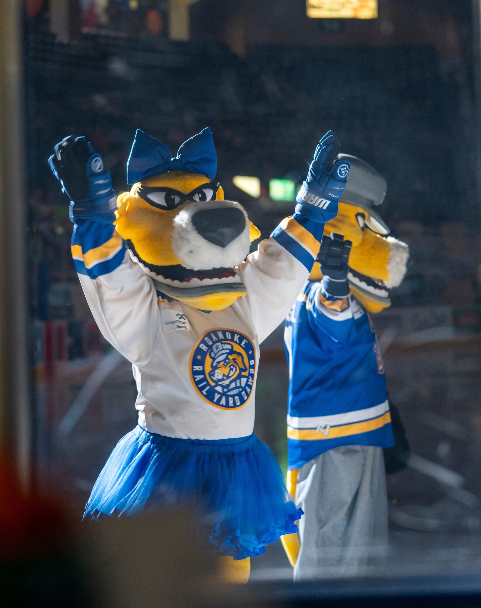 Two mascot characters in hockey jerseys and costumes, one with a blue tutu, celebrating with raised arms in an arena.