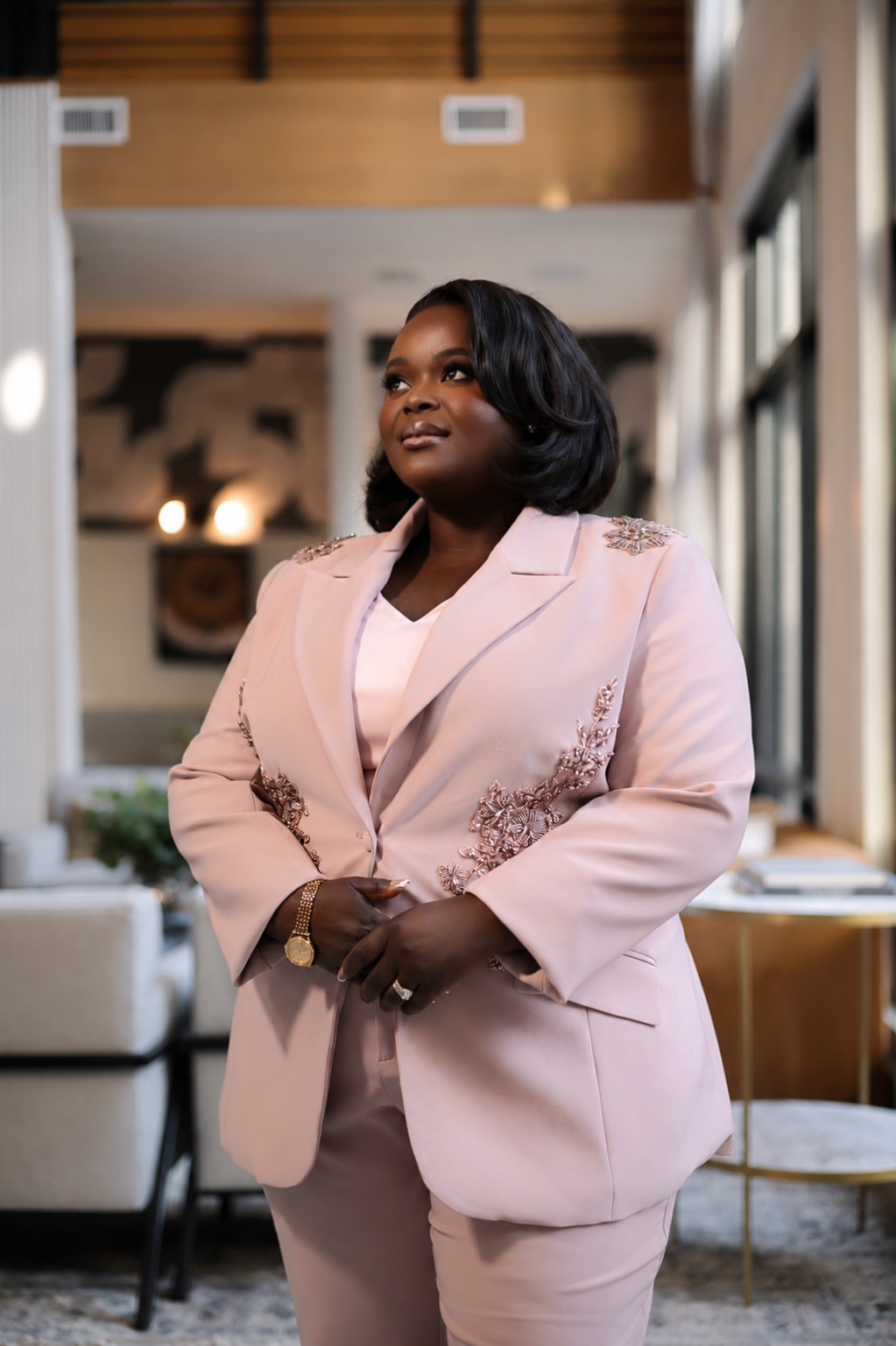 Woman in pink suit standing indoors with large windows and modern decor, smiling confidently.