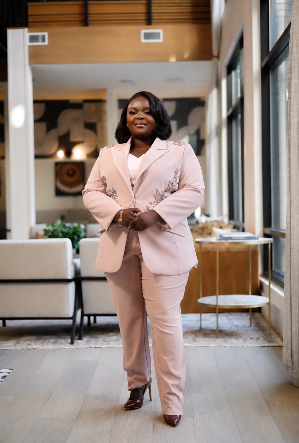 Woman in pink suit standing in a modern, well-lit indoor space with large windows and furniture.