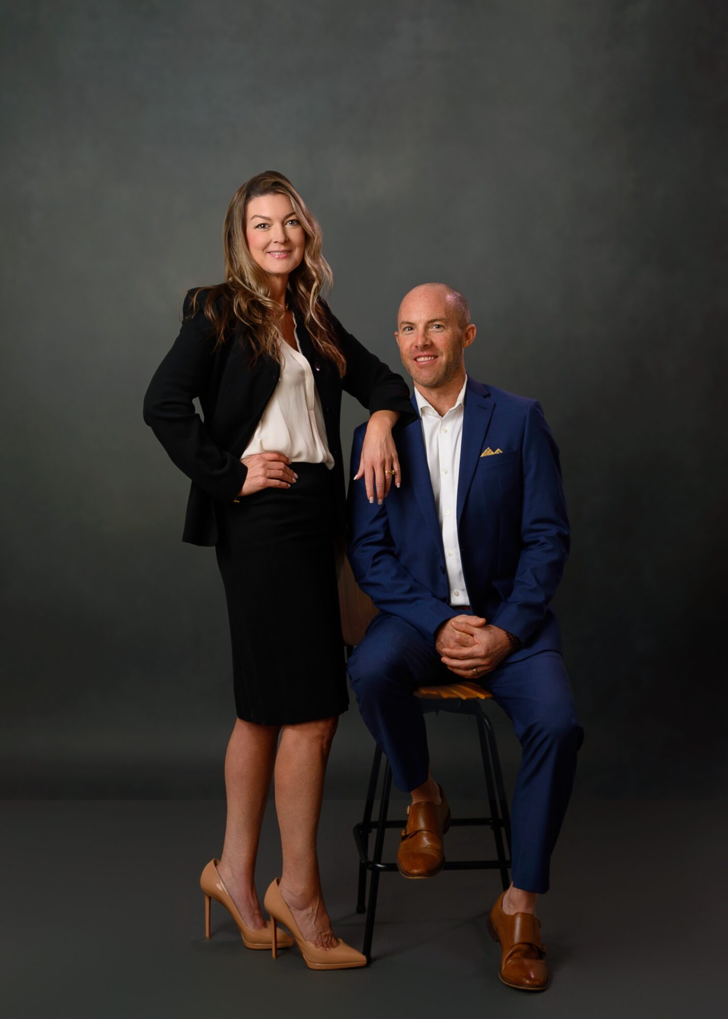 A woman stands next to a seated man against a plain dark background. She is smiling, wearing a black blazer, skirt, and beige heels. The man is in a blue suit, white shirt, and brown shoes, smiling and looking at the camera.