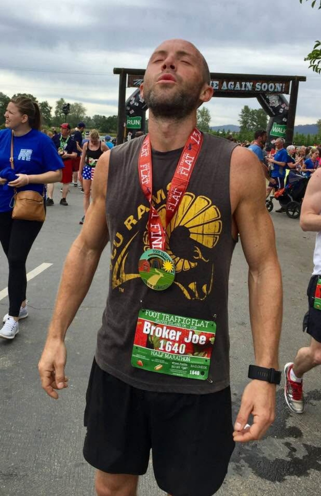 Man with a beard and shaved head wearing a race bib and medal, standing outdoors after a race, with other runners in background.
