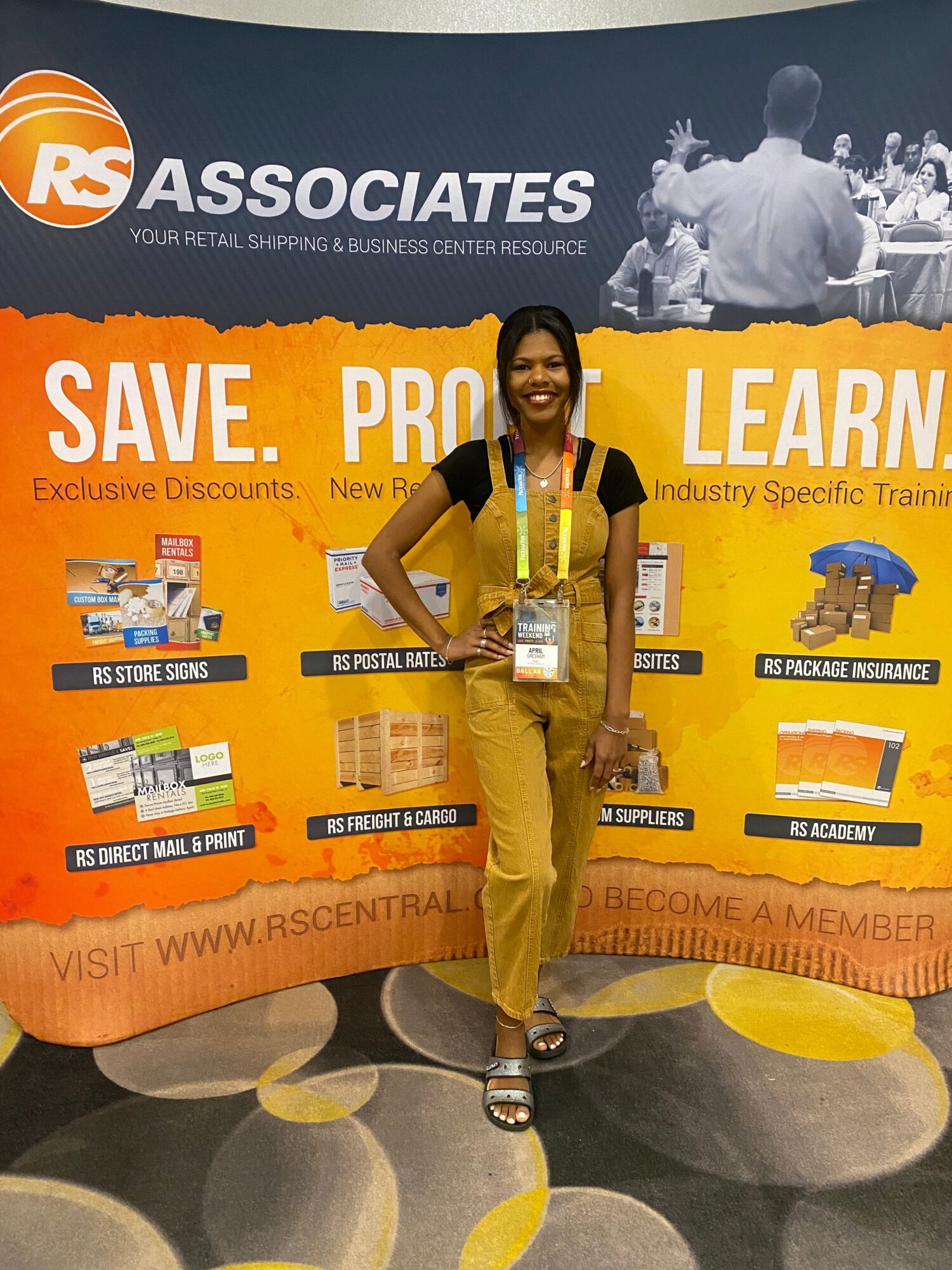 Person standing in front of a large orange and yellow promotional banner, wearing yellow pants and a black top, holding a camera.