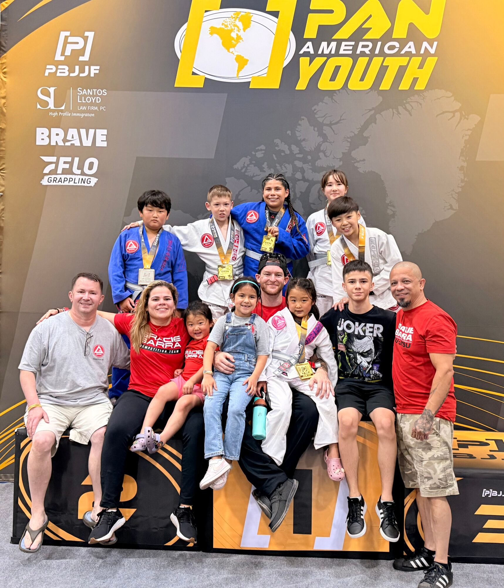 Group of young athletes and coaches posing on a podium at a martial arts event, with medals and a banner in the background.