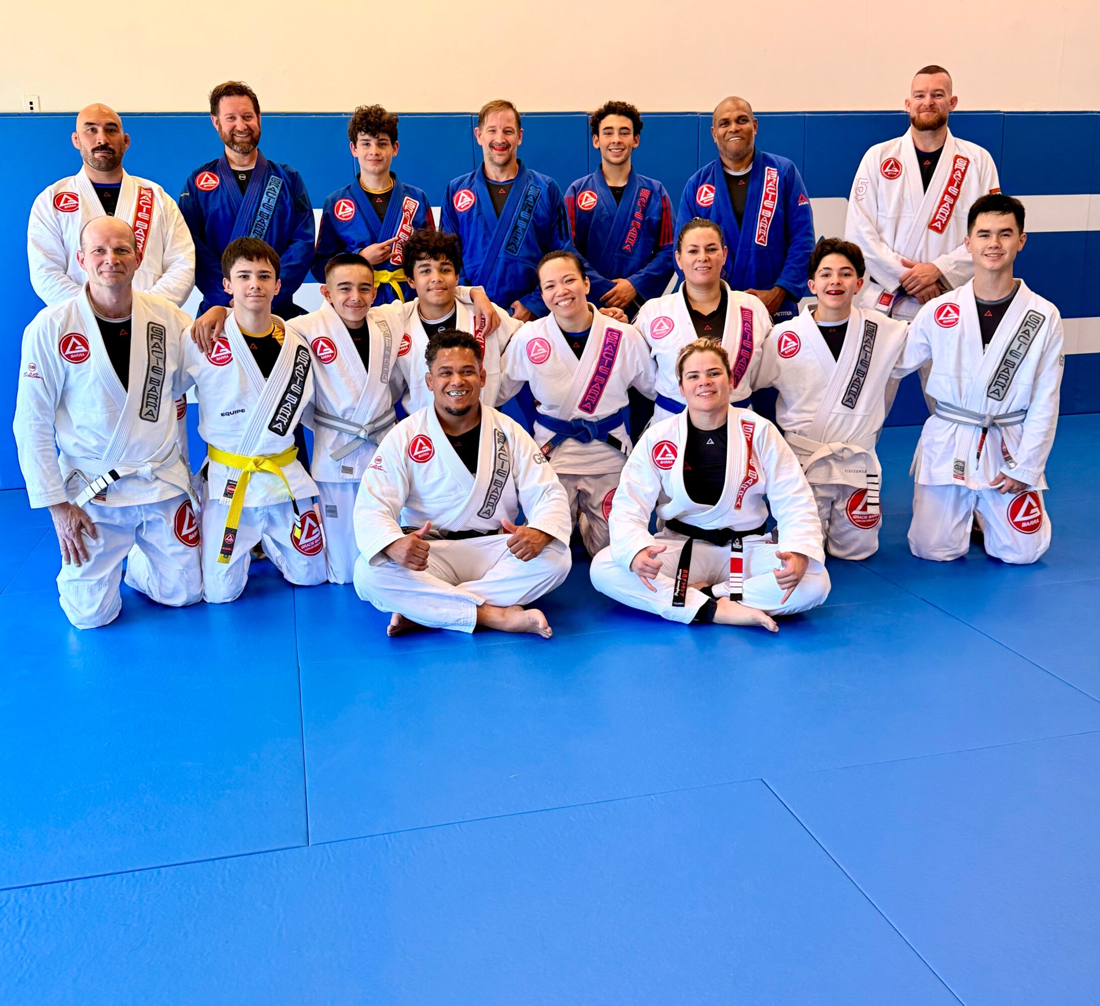 Group of people in Brazilian Jiu-Jitsu gis posing on blue mat, some kneeling and some standing, in a training facility.