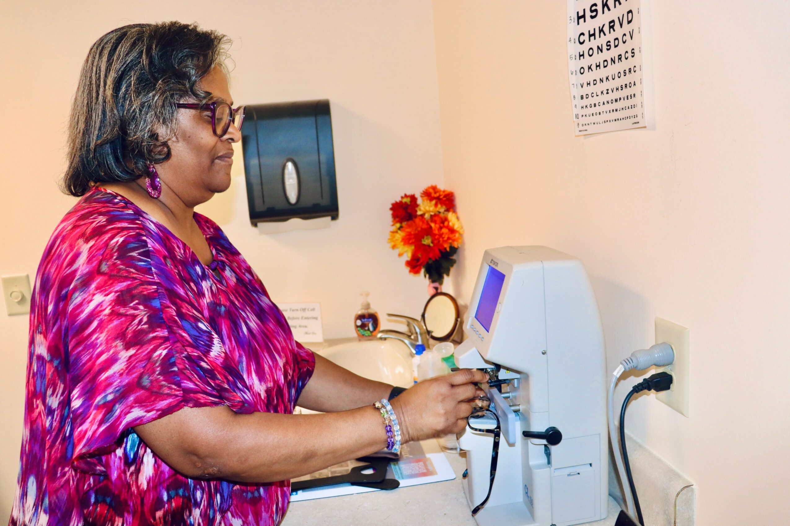 Woman in colorful blouse using medical device in clinic, eye chart on wall, flowers on counter, paper on table