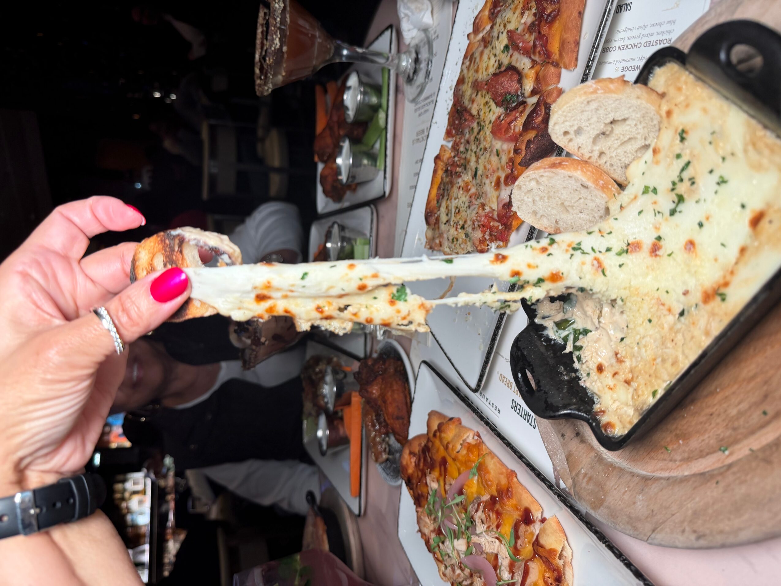 Person holding a slice of cheesy baked dish with bread, with pizza and other dishes on table in background.