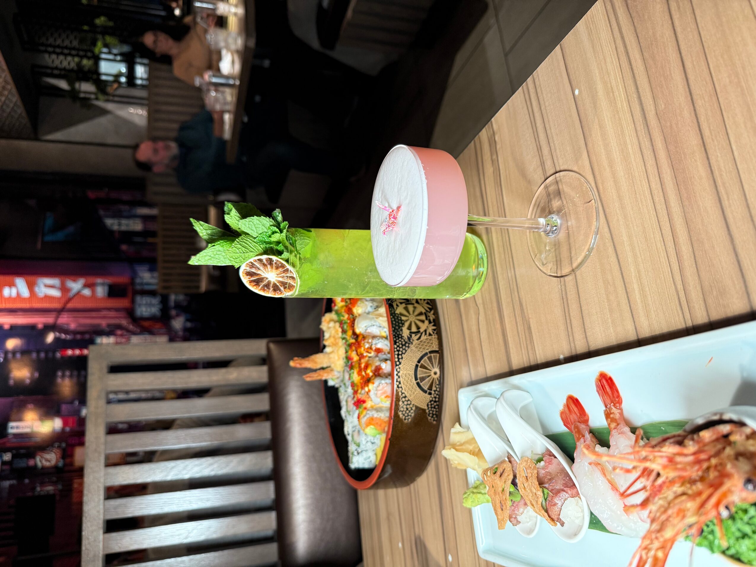 A green cocktail with garnishes, a sushi platter, and a pink drink on a wooden table in a restaurant.