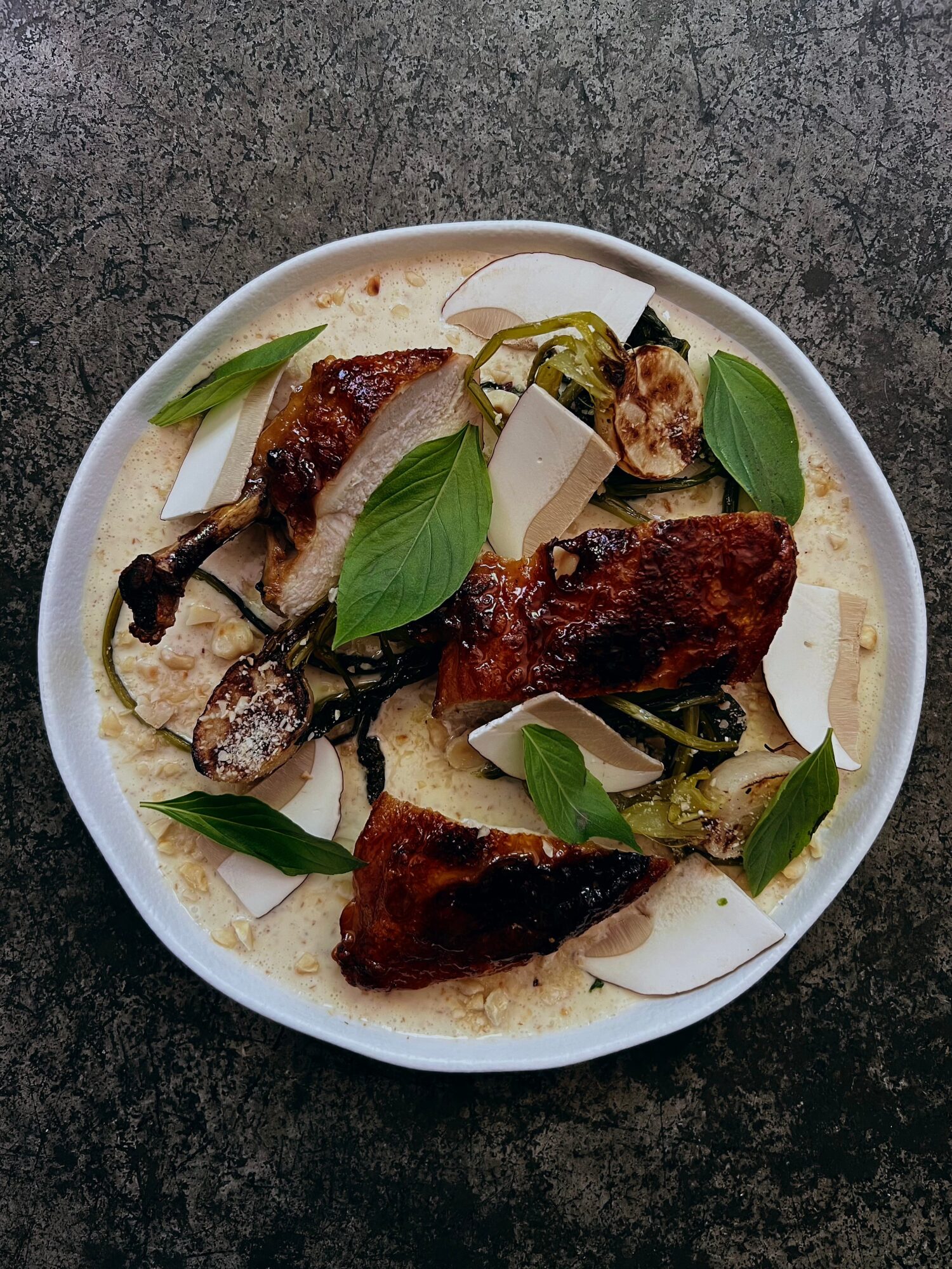 Roasted chicken pieces with green leaves and cheese slices on a white plate, on a dark surface.
