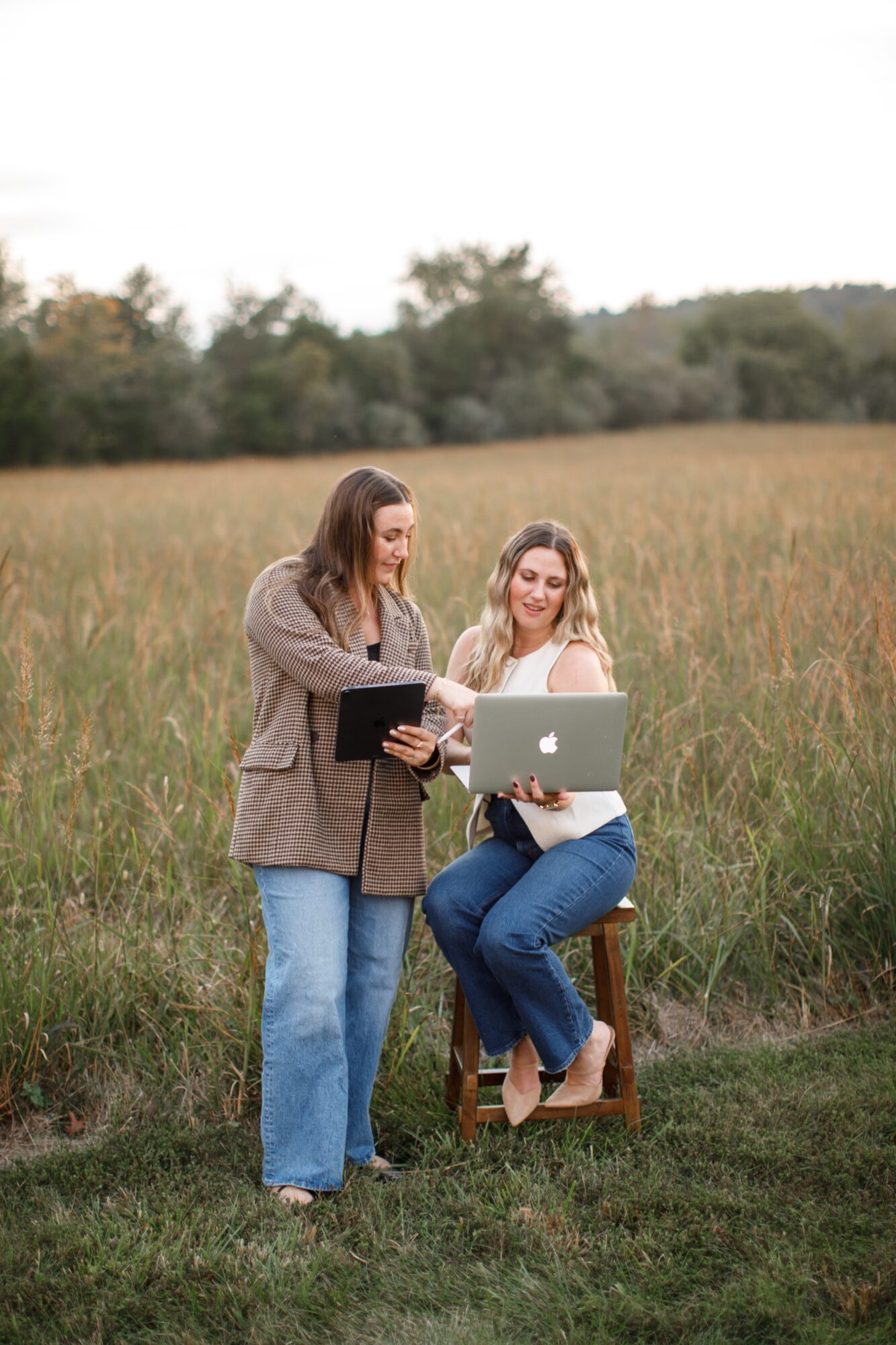 Two women outdoors in a field, one sitting on a stool with a laptop, the other standing with a tablet, smiling.