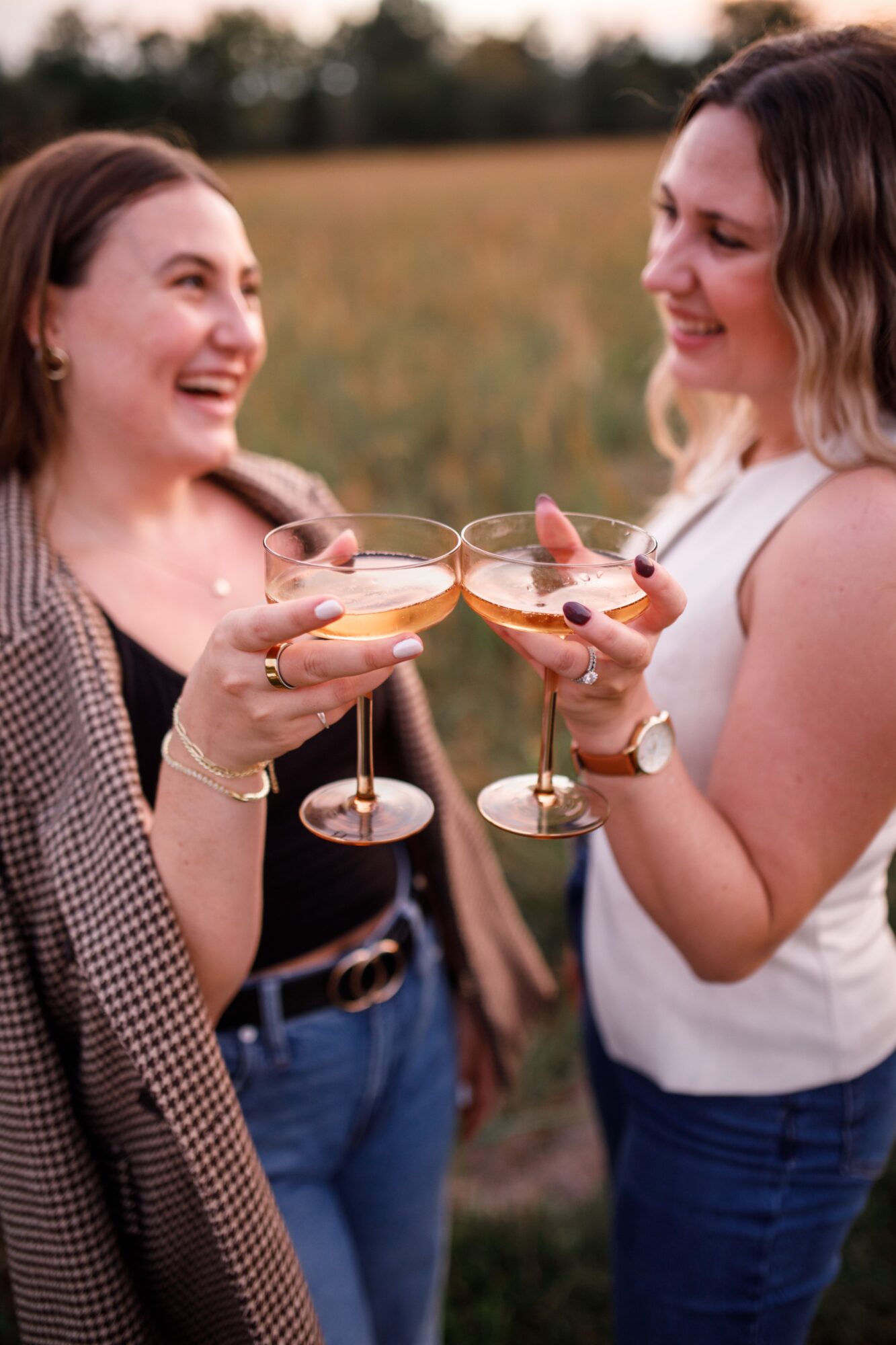 Two women smiling and clinking glasses of champagne outdoors in a field, enjoying a celebration.