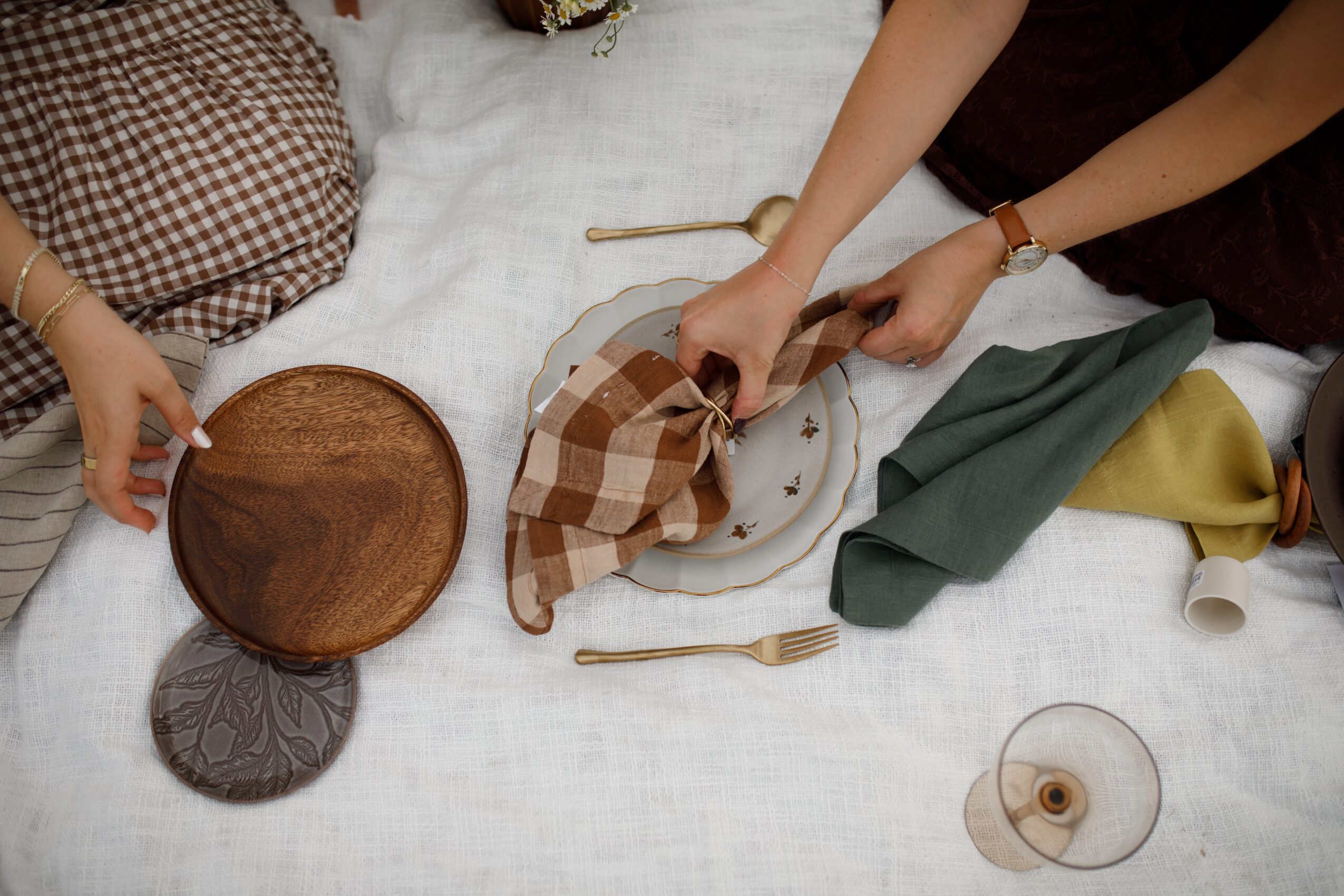 People setting a table with plates, napkins, and utensils on a white tablecloth, with hands arranging items.