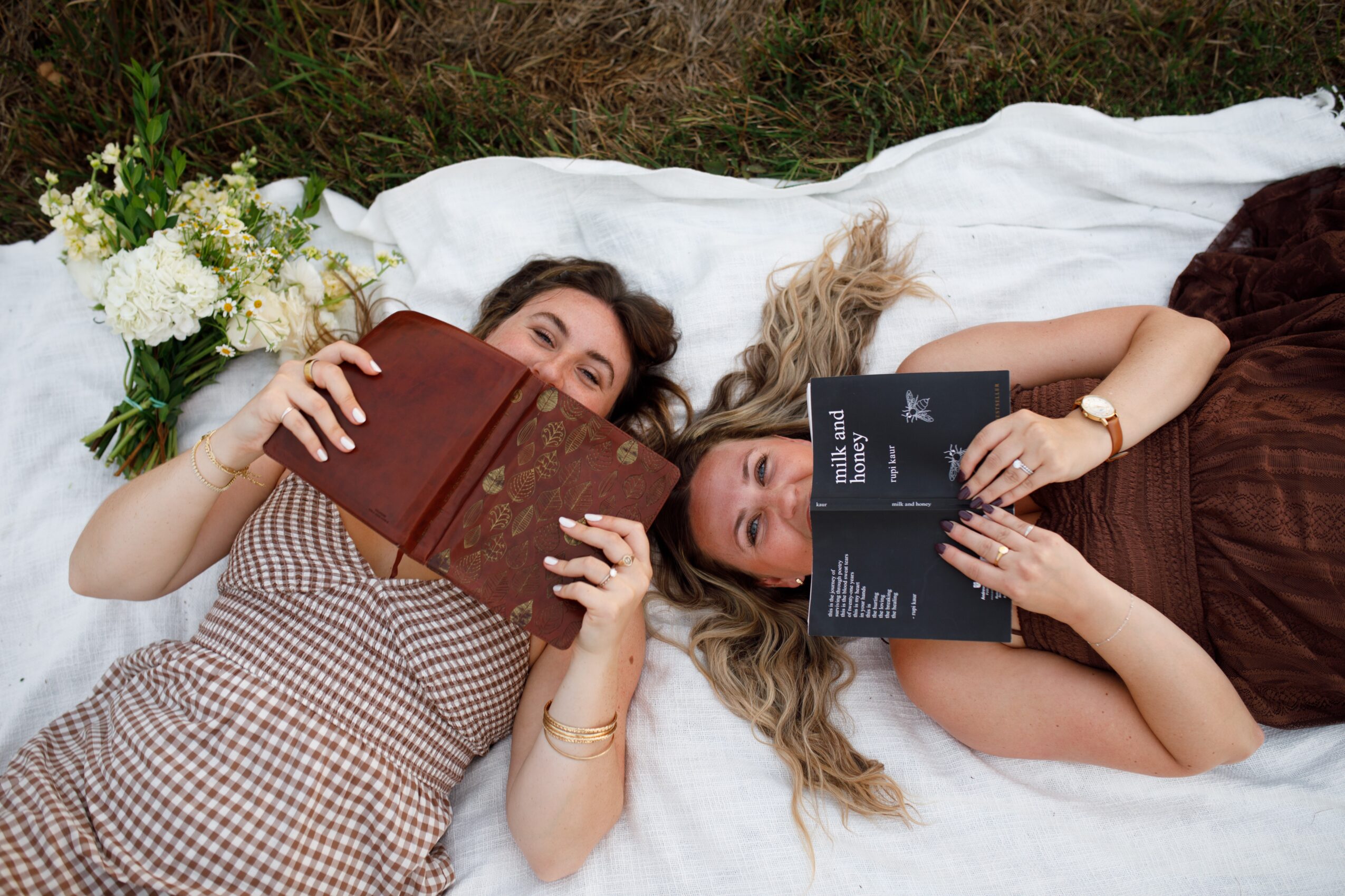 Two women lie on a bed, reading books, with flowers nearby, one with dark hair and the other with blonde hair.