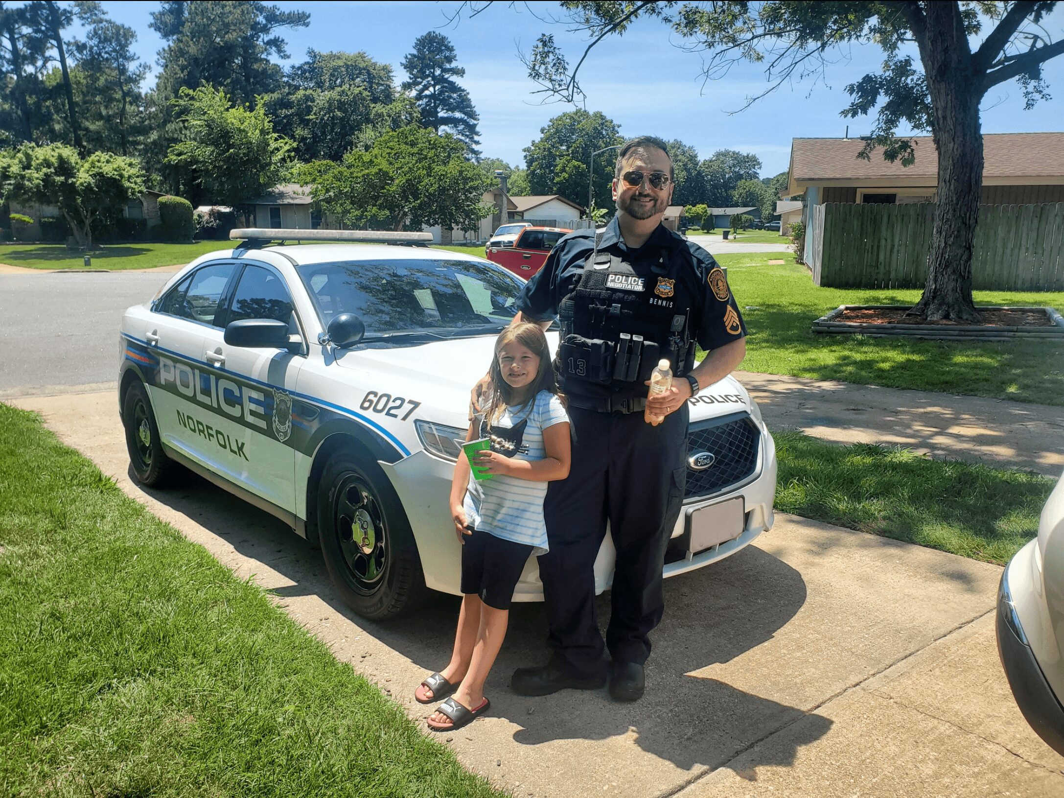 A police officer and a young girl stand together on a sidewalk next to a police car in a residential neighborhood on a sunny day.