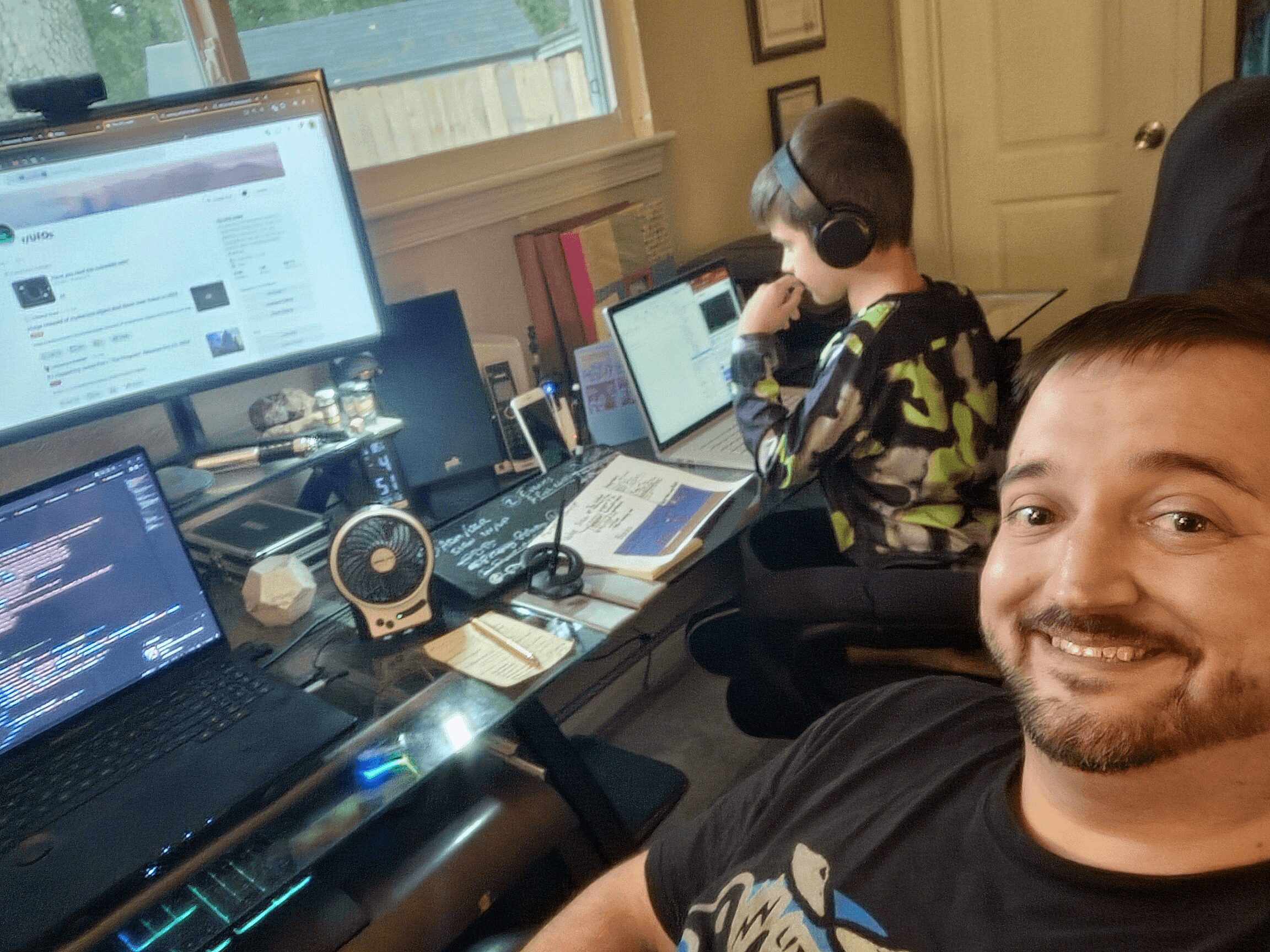 Two people working at desks with multiple computer screens, one smiling at the camera, the other wearing headphones and focused on a laptop.